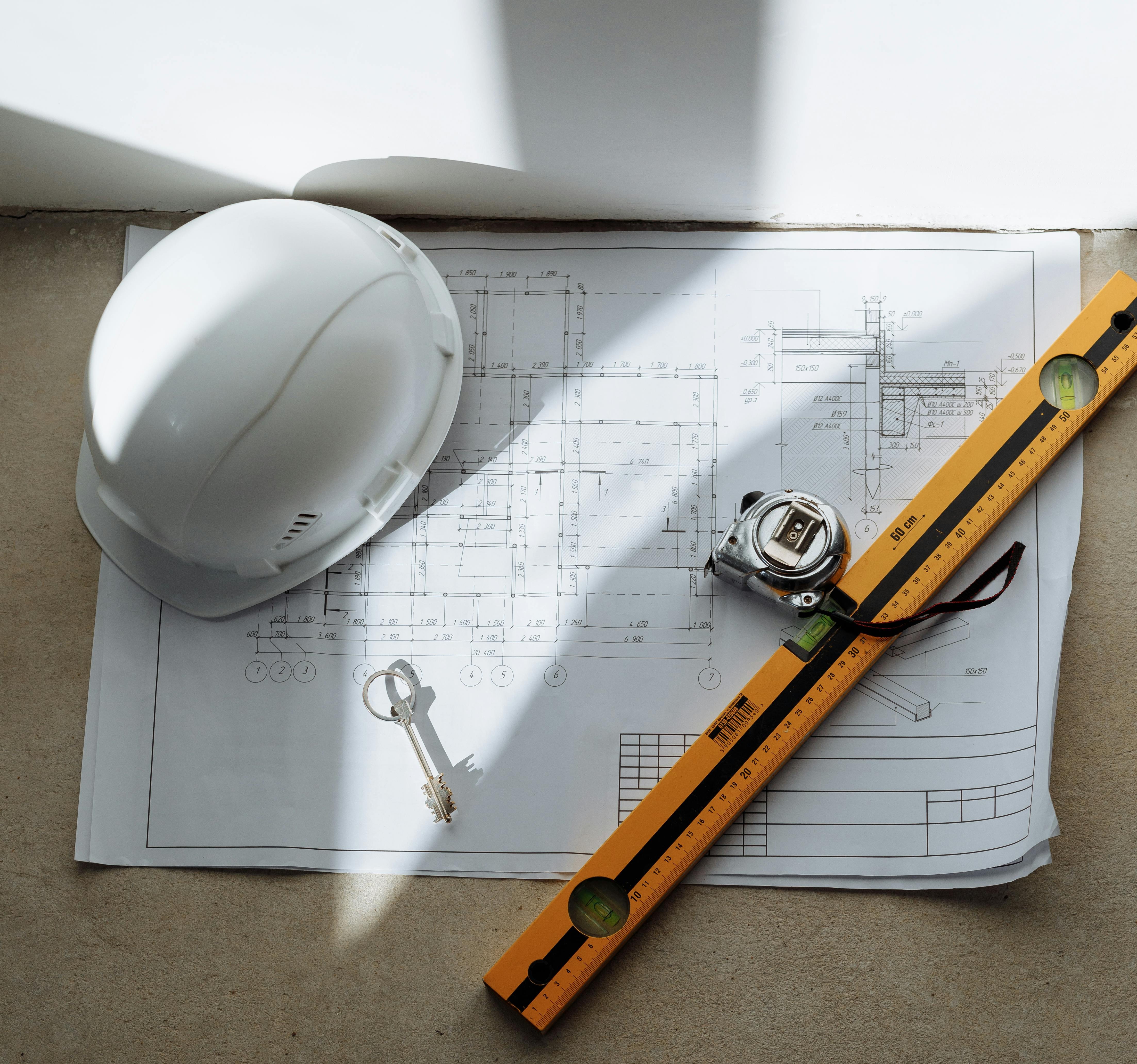 Professional construction planning tools including blueprints, hard hat, level, measuring tape, and keys on concrete surface